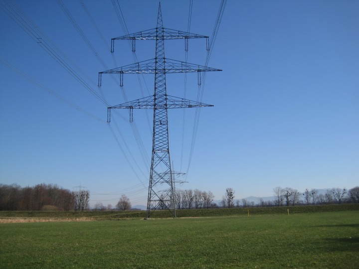 Das Bild zeigt einen einzelnen mast für Hochspannungsleitunge. Er steht in einer grünen, offenen Landschaft, wahrscheinlich ein Feld oder eine Wiese.
Leitungen:
Vom Mast aus erstrecken sich mehrere Hochspannungsleitungen in verschiedene Richtungen. Sie verlaufen in einem leichten Durchhang zwischen den Masten, die außerhalb des Bildausschnitts liegen.
Umgebung:
Der Mast steht auf einer grünen Fläche mit kurzem Gras. Im Hintergrund ist eine Baumreihe zu sehen, die den Horizont bildet. Der Himmel ist klar und blau, ohne Wolken. Das Licht deutet auf einen sonnigen Tag hin.