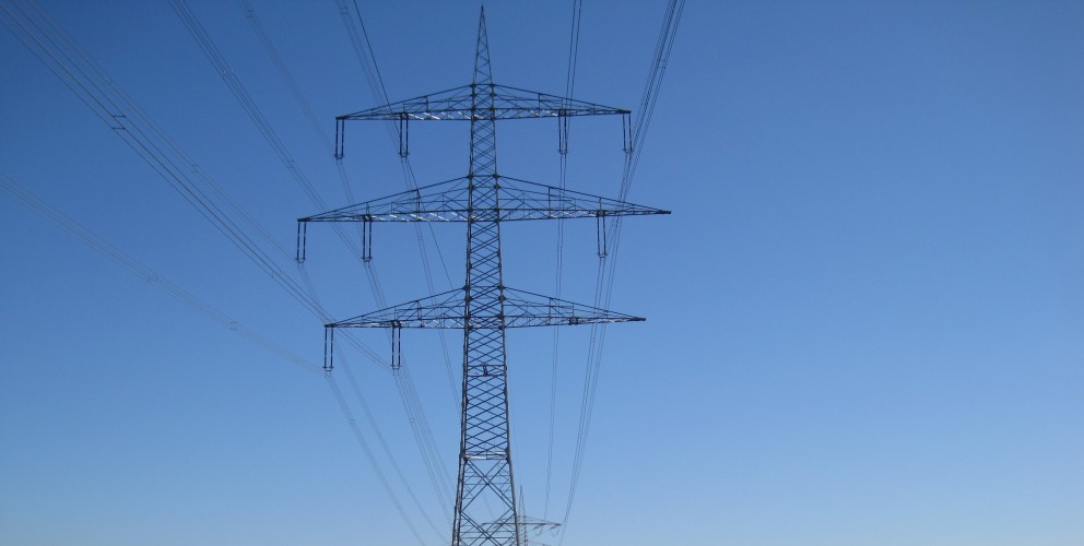 Das Bild zeigt einen einzelnen mast für Hochspannungsleitunge. Er steht in einer grünen, offenen Landschaft, wahrscheinlich ein Feld oder eine Wiese.
Leitungen:
Vom Mast aus erstrecken sich mehrere Hochspannungsleitungen in verschiedene Richtungen. Sie verlaufen in einem leichten Durchhang zwischen den Masten, die außerhalb des Bildausschnitts liegen.
Umgebung:
Der Mast steht auf einer grünen Fläche mit kurzem Gras. Im Hintergrund ist eine Baumreihe zu sehen, die den Horizont bildet. Der Himmel ist klar und blau, ohne Wolken. Das Licht deutet auf einen sonnigen Tag hin.