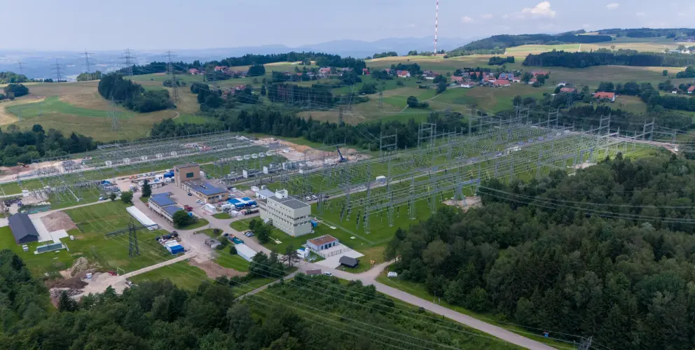 Luftaufnahme der Umspannanlage Kühmoos, eines großen Stromnetzknotens im ländlichen Südschwarzwald. Die weitläufige Anlage nimmt den unteren und mittleren Teil des Bildes ein und besteht aus mehreren Betriebsgebäuden und einem komplexen System aus Freiluftschaltanlagen, Transformatoren und zahlreichen Hochspannungsleitungen und Masten. 