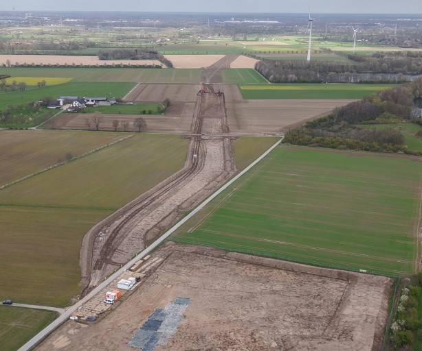 Luftaufnahme einer linienförmigen Baustelle (Trassenbau) inmitten von Feldern und Wiesen. Der Erdaushub zieht sich als brauner Streifen von einer großen Baufläche im Vordergrund weit nach hinten bis zum Horizont.