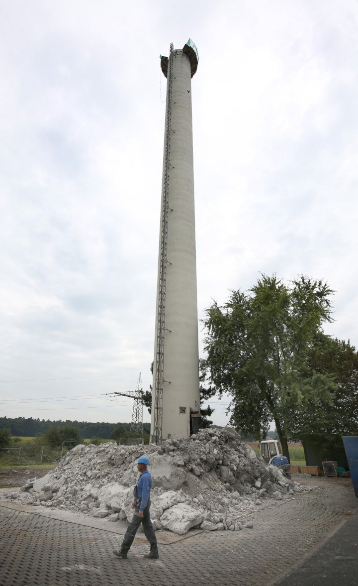 Es folgt eine Bildbeschreibung:
Das Foto im Hochformat zeigt einen halb abgerissenen Richtfunkturm. Der obere Teil ist abgerissen, am Fuße des Turm liegt Schutt. Im Vordergrund passiert ein Arbeiter die Baustelle.
Ende der Bildbeschreibung.
