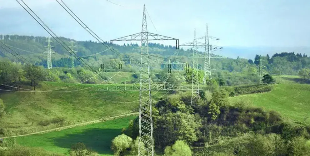 Eine weite Landschaftsansicht mit mehreren großen Stahlfachwerk-Strommasten, die Hochspannungsleitungen über hügelige, grüne Wiesen und bewaldete Hänge spannen. 
