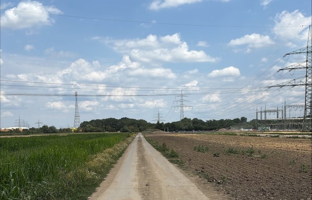 Fotografie einer ländlichen Ebene mit Feldern, durch die ein Feldweg führt. Im Hintergrund und am rechten Bildrand sind mehrere Hochspannungsmasten und ein Umspannwerk unter einem leicht bewölkten Himmel zu sehen.