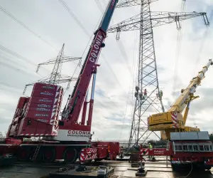 Zwei große Kräne heben Arbeiter an einem Hochspannungsmast mit mehreren Stromleitungen