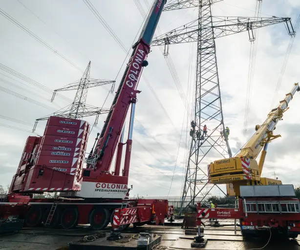 Zwei große Kräne heben Arbeiter an einem Hochspannungsmast mit mehreren Stromleitungen