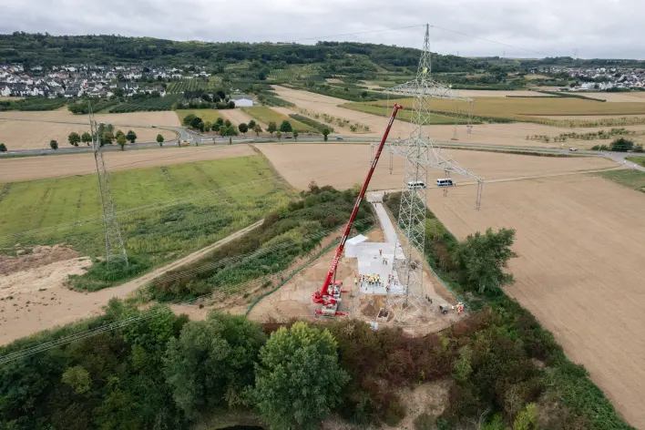 Drohnenaufnahme einer Mastbaustelle: Ein neuer Hochspannungsmast wird mit einem roten Kran montiert. Im Hintergrund sind Felder, eine Ortschaft und weitere Masten zu sehen.