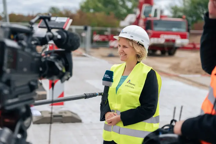 Eine Frau mit weissem Amprion-Schutzhelm und gelber Warnweste gibt dem Kamerateam von SWR Aktuell ein Interview auf einer Hochspannungsbaustelle. Im Hintergrund sind ein roter Kran und Baufahrzeuge zu sehen.