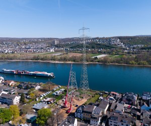 Luftaufnahme eines Flusses mit einem großen Strommast, einem Kran und einem Frachtschiff sowie Wohnhäusern am Ufer