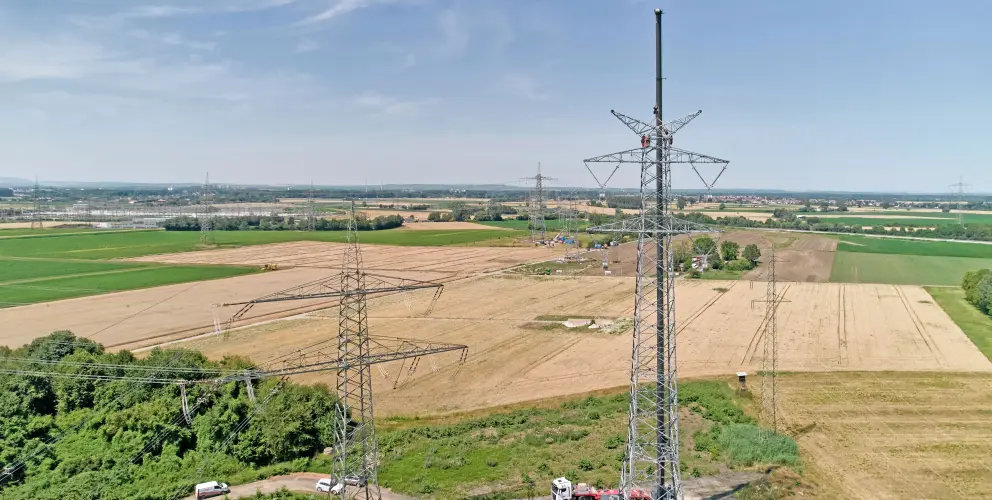 Vordergrund und Mitte: Das Bild wird dominiert von einem großen, Gittermast einer Hochspannungsleitung, der sich bis in den oberen rechten Bereich erstreckt. Er hat komplexe Isolatorstrukturen und eine Antenne oder ähnliches an der Spitze. Darunter liegen landwirtschaftliche Flächen – einige Felder sind grün, andere braun oder gelb. Ein kleinerer, ähnlicher Mast ist links unten teilweise sichtbar.
Hintergrund: Weitere Masten der Stromleitungen sind in der Ferne über die weite, flache Landschaft verteilt. Am Horizont ist eine Ansammlung von Gebäuden und Industriebauten, möglicherweise eine kleine Stadt oder ein Gewerbegebiet, schwach erkennbar. Dahinter erstrecken sich sanfte Hügel oder Berge unter einem hellblauen Himmel mit einigen weißen Wolken.
