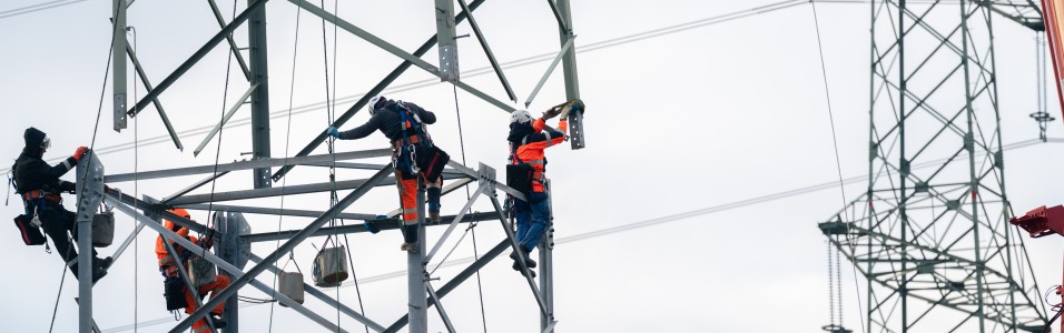 Mehrere Arbeiter in Schutzkleidung klettern an einem hohen Stahlgittermast für Stromleitungen