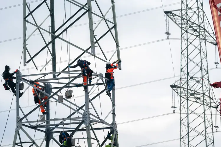 Mehrere Arbeiter in Schutzkleidung klettern an einem hohen Stahlgittermast für Stromleitungen