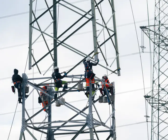 Vier Monteure in oranger Schutzkleidung und Helmen arbeiten gesichert an der Stahlkonstruktion eines Hochspannungsmastes in großer Höhe. Im Hintergrund ist ein weiterer Mast zu sehen.
