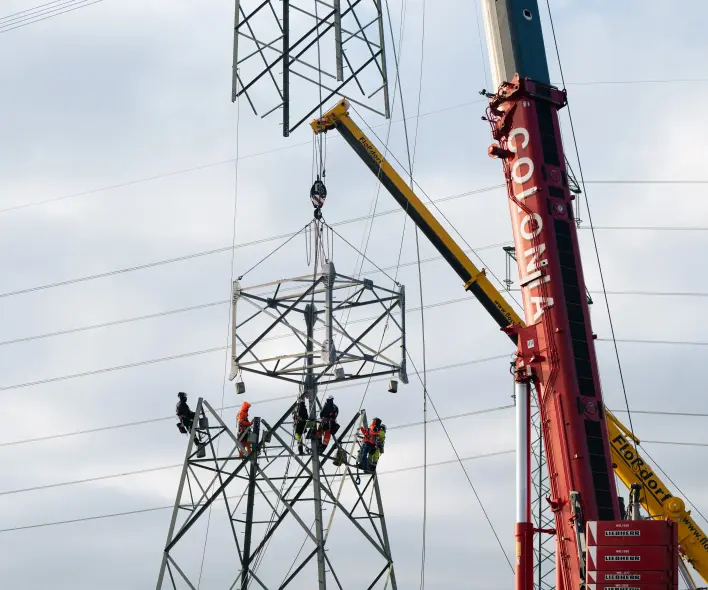 Ein Kran hebt ein Stahlsegment (Zwischenschuss) zu einem Hochspannungsmast. Mehrere gesicherte Monteure stehen auf dem Mast und begleiten die Montage. Im Hintergrund sind Stromleitungen zu sehen.