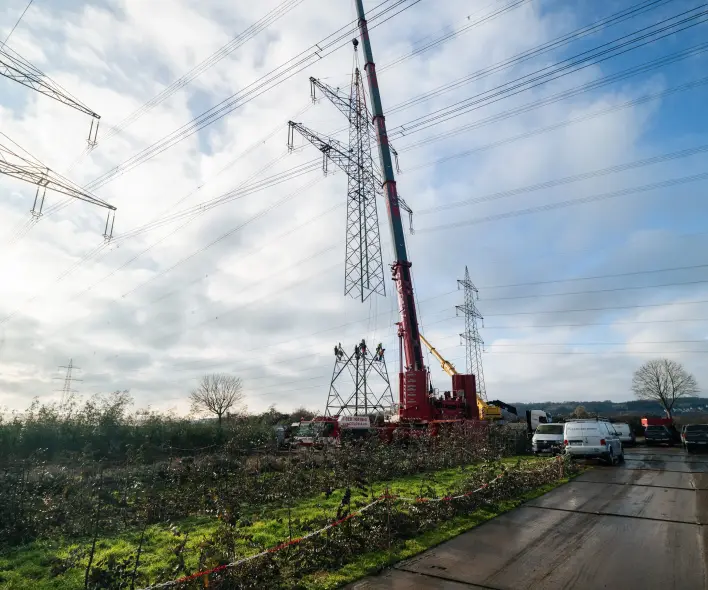 Seitenansicht einer Masterhöhungs-Baustelle: Ein roter Kran steht neben einem im Aufbau befindlichen Hochspannungsmast. Links und rechts sind bestehende Masten mit Stromleitungen zu sehen. Im Vordergrund verläuft eine Feldstraße.
