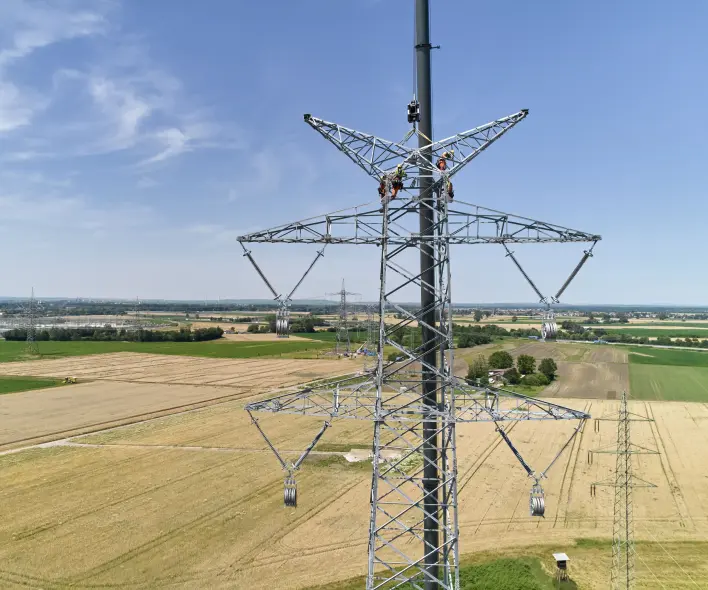 Drohnenaufnahme eines im Bau befindlichen Hochspannungsmastes bei Lampertheim/Bürstadt: Zwei Monteure arbeiten in großer Höhe an der Mastspitze. Im Hintergrund erstreckt sich eine flache Agrarlandschaft mit Feldern und weiteren Masten.