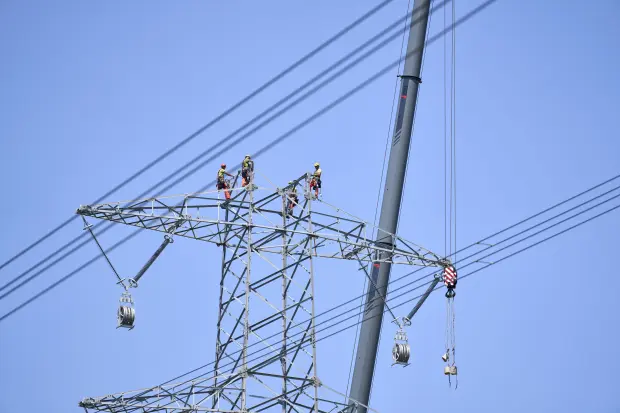 Drei Monteure in Schutzkleidung und Helmen arbeiten gesichert auf dem Querausleger eines Hochspannungsmastes in großer Höhe. Neben ihnen ist der Arm eines Krans zu sehen. Im Hintergrund ein wolkenloser blauer Himmel.
