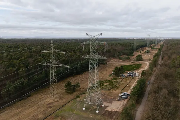 Drohnenaufnahme zweier Hochspannungsmasten in einer Waldschneise: Am rechten Mast befinden sich Baufahrzeuge und Geräte am Boden. Die bewaldete Trasse erstreckt sich in die Ferne. Am Horizont ist eine Stadtsilhouette zu erkennen.