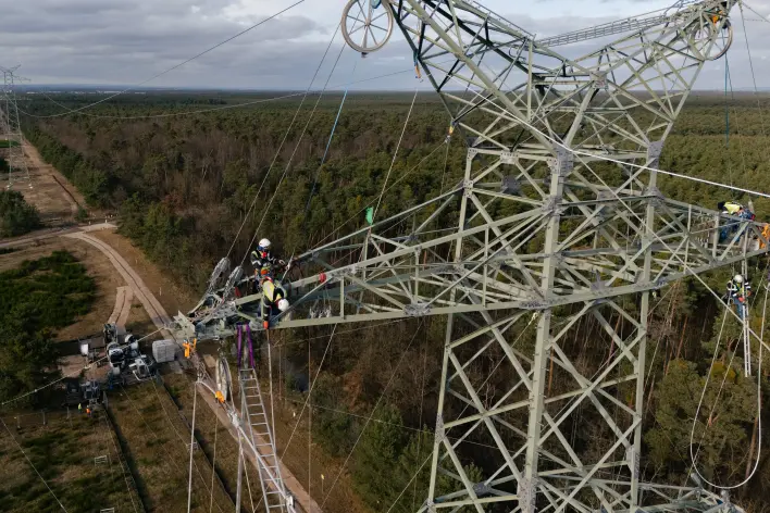 Drohnennahaufnahme des Querauslegers eines Hochspannungsmastes in einer Waldschneise: Mehrere Monteure in Schutzkleidung und Helmen arbeiten gesichert auf dem Stahlträger. Im Hintergrund erstreckt sich die bewaldete Trasse bis zum Horizont.