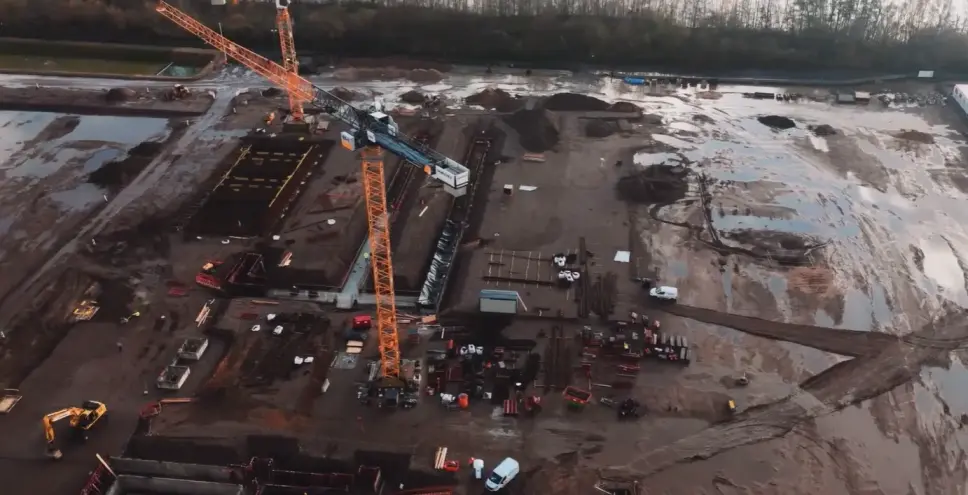 Luftaufnahme der Ultranet-Konverterstandort-Baustelle in der Hochbauphase: orangefarbener Turmdrehkran, Fundamentstrukturen und Baufahrzeuge auf weitläufigem Baufeld.
