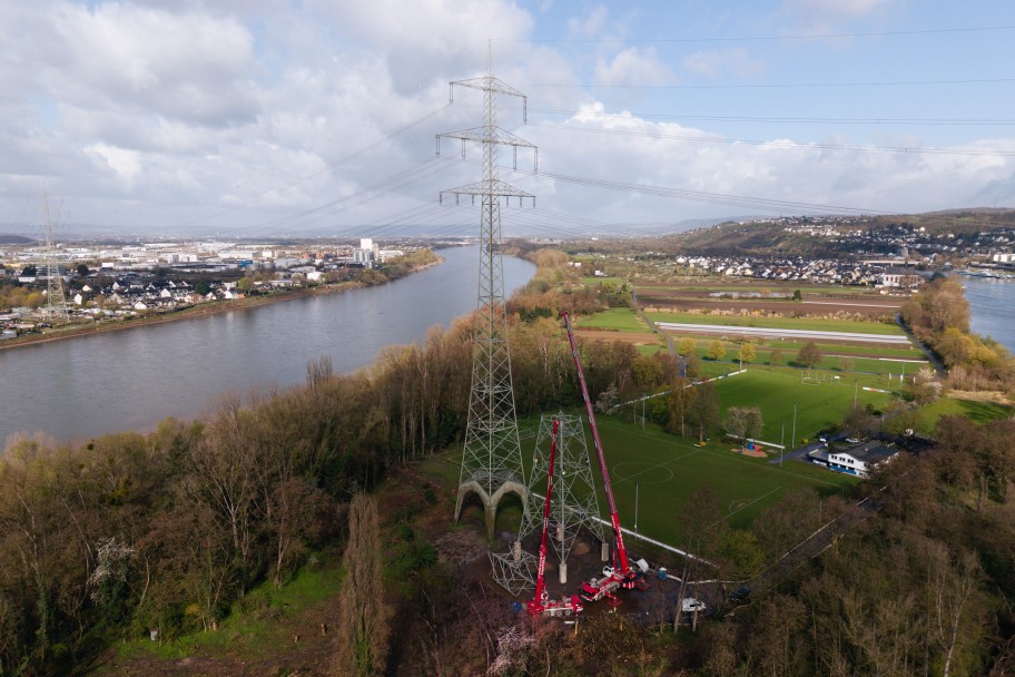 Luftaufnahme eines Flusses mit Hochspannungsmast und Baukränen in einer grünen Landschaft nahe einer Stadt
