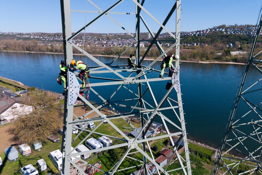 Drei Personen in Schutzkleidung klettern an einem hohen Stahlgittermast über einem Fluss