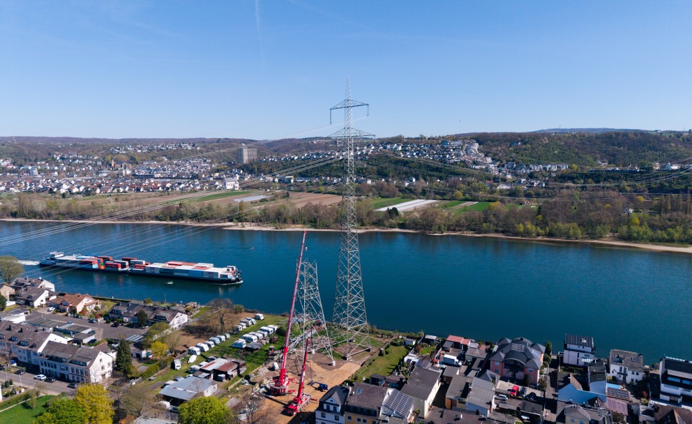 Luftaufnahme eines Flusses mit einem großen Strommast, einem Kran und einem Frachtschiff sowie Wohnhäusern am Ufer