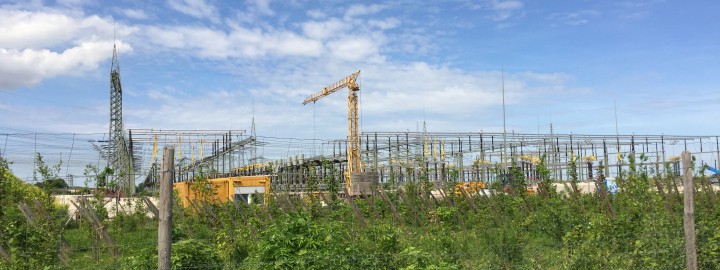 Blick auf eine große Umspannstation im Bau, aufgenommen bei Tageslicht. Im Vordergrund ein grüner Zaun mit wuchernder Vegetation. Dahinter zahlreiche Stahlgerüste, Transformatoren und Leitungen, die über die gesamte Breite verlaufen. Zwei gelbe Baukräne ragen hoch auf, einer in der Mitte und einer weiter links. Im Hintergrund sieht man einen blau-weißen Himmel mit vereinzelten Wolken.