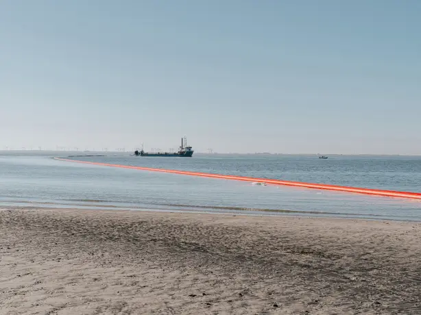 Das Bild zeigt maritime Bauarbeiten an der Küste, bei denen eine auffällige, hellrote Leitung vom Meer an den Strand geführt wird. Diese Art von Leitung, oft mit Auftriebskörpern versehen, wird üblicherweise für Offshore-Windparkprojekte oder Küstenrenaturierungsmaßnahmen wie das Aufspülen von Sand verwendet. 