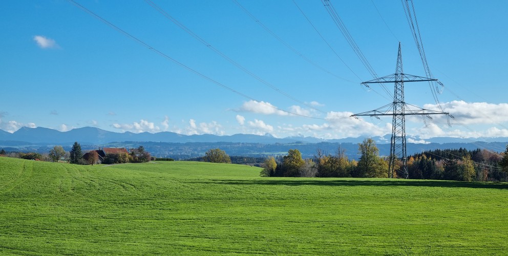 Eine weite, hellgrüne Wiese unter einem blauen Himmel mit weißen Wolken. Im mittleren Bereich steht ein großer Strommast aus Stahl, der mehrere Hochspannungsleitungen über das Feld führt. Im Hintergrund sind dunkle Berge oder Hügel am Horizont zu sehen. Rechts und links vom Feld stehen Bäume in herbstlichen Farben.