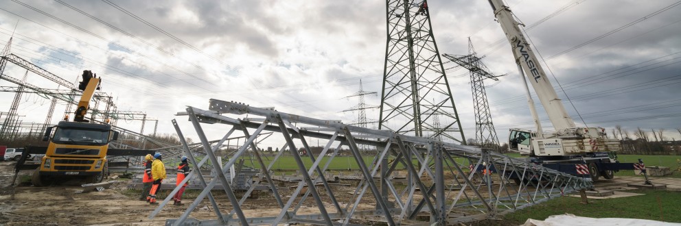 Das Bild zeigt eine Baustelle, auf der Arbeiten an einem oder mehreren großen Strommasten durchgeführt werden, möglicherweise ein "Mastkopfwechsel", wie im Titel angedeutet.
Im Vordergrund liegen mehrere Elemente einer Gittermastkonstruktion auf dem Boden. Diese scheinen neu zu sein oder demontiert worden zu sein.
In der Mitte des Bildes steht ein hoher, schlanker Strommast. An diesem Mast ist ein großer, weißer Mobilkran mit einem langen Ausleger positioniert. Der Ausleger reicht hoch zum Kopf des Mastes. Es ist wahrscheinlich, dass der Kran verwendet wird, um schwere Teile des Mastkopfes auszutauschen oder zu montieren.
Links im Bild ist ein gelb-schwarzer LKW-Kran mit einem ebenfalls ausgefahrenen Arm zu sehen, der sich in der Nähe weiterer Gittermaststrukturen befindet. Mehrere Bauarbeiter in orangefarbener Warnkleidung und gelben Helmen bewegen sich in der Nähe der Baustelle.
Im Hintergrund sind weitere Strommasten und Hochspannungsleitungen zu erkennen, die sich durch eine offene Landschaft ziehen. Der Himmel ist bedeckt mit grauen und weißen Wolken, was auf einen trüben Tag hindeutet.