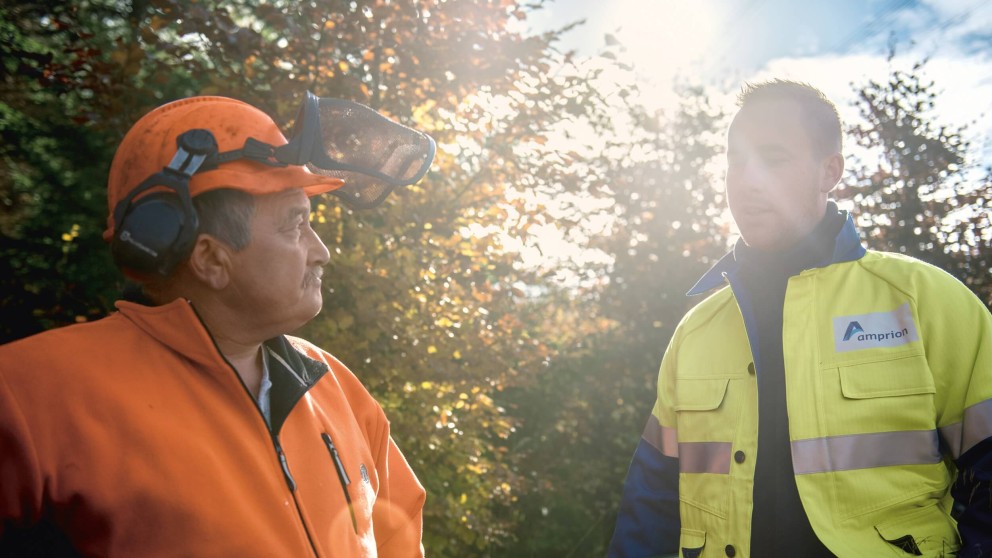 Es folgt eine Bildbeschreibung:
Die Fotografie im Querformat zeigt zwei Herren vor Bäumen, die Sonne scheint zwischen den Bäumen hindurch. Sie sind einander zugewandt und augenscheinlich im Gespräch. Der linke Herr trägt orangefarbene Bekleidung, einen orangenen Helm mit Gehör – und Augenschutz. Der Mann hat einen Schnauzbart und hat die Hände in die Hüften gesenkt. Der Herr in der rechten Bildhälfte trägt eine neon-gelbe Sicherheitsjacke, welche blau abgesetzt ist. Auf seiner Brust ist – etwa postkartengroß – das Logo der Amprion-GmbH aufgedruckt. Der Herr trägt kurzes Haar und einen gepflegten Drei-Tage-Bart.
Ende der Bildbeschreibung