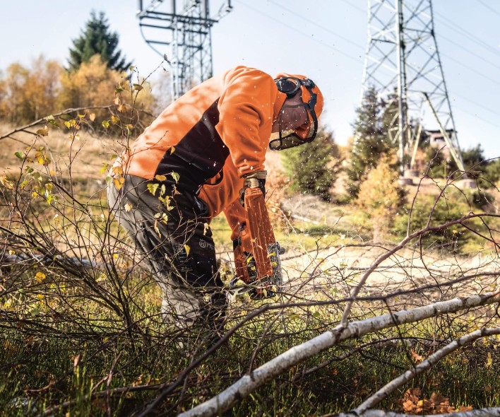 Es folgt eine Bildbeschreibung:
Die Fotografie im Querformat zeigt Rückschnittarbeiten an einem leicht bewaldeten Hang. Im Zentrum des Bildes ist ein Arbeiter in orangefarbener Arbeitskleidung zu sehen. Der Arbeiter trägt einen orangenen Helm mit Gehör- und Augenschutz und hält in der rechten Hand eine benzinbetriebene Kettensäge. Er ist nach vorne gelehnt und sägt gerade einen Ast durch. Im Hintergrund, leicht unscharf sind die unteren Teile eines Gittersmasten zu sehen, vor dem Arbeiter bereits abgesägte Teile von Bäumen. Das Bild erzeugt eine professionelle und nicht unfreundliche Emotion.
Ende der Bildbeschreibung.