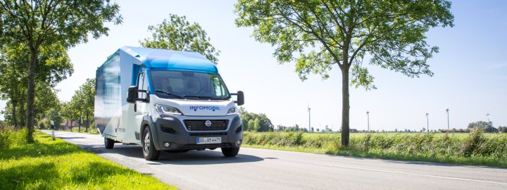 A-Nord vor Ort. Das Foto zeigt das blau-lilane Infomobil im Sonnenschein während der Fahrt auf einer asphaltierten Landstraße/Allee. 