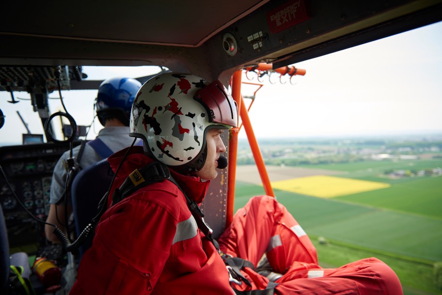 Das Bild zeigt einen Monteur in rotem Overall. Er sitzt in einem fliegenden Helikopter mit den Füßen außerhalb der Tür.