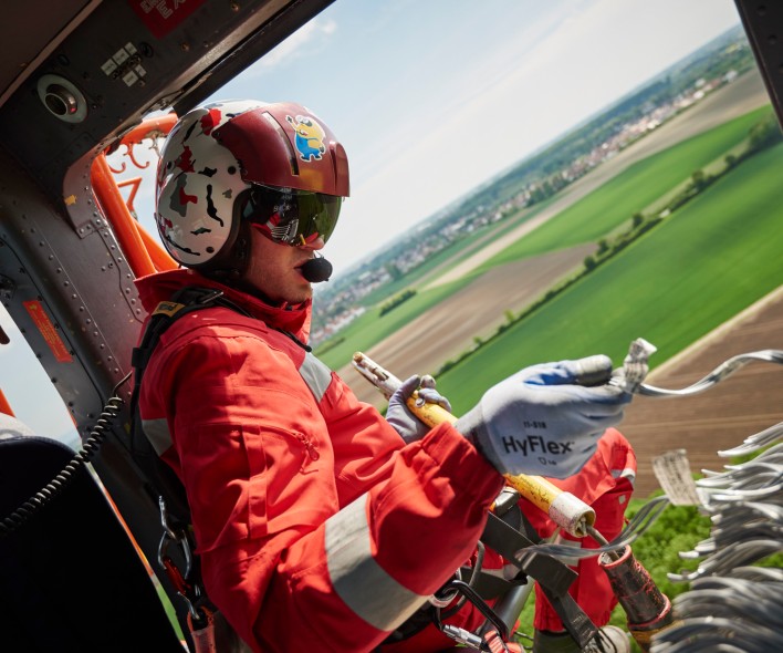 Das Bild zeigt einen Monteur in rotem Overall. Er sitzt in einem fliegenden Helikopter mit den Füßen außerhalb der Tür und montiert an den Trassen.