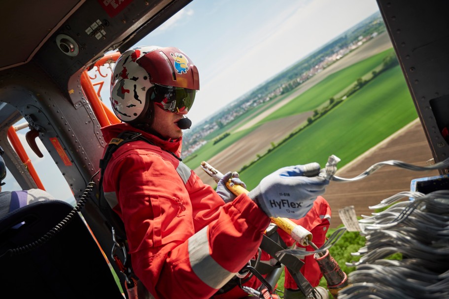 Das Bild zeigt einen Monteur in rotem Overall. Er sitzt in einem fliegenden Helikopter mit den Füßen außerhalb der Tür und montiert an den Trassen.