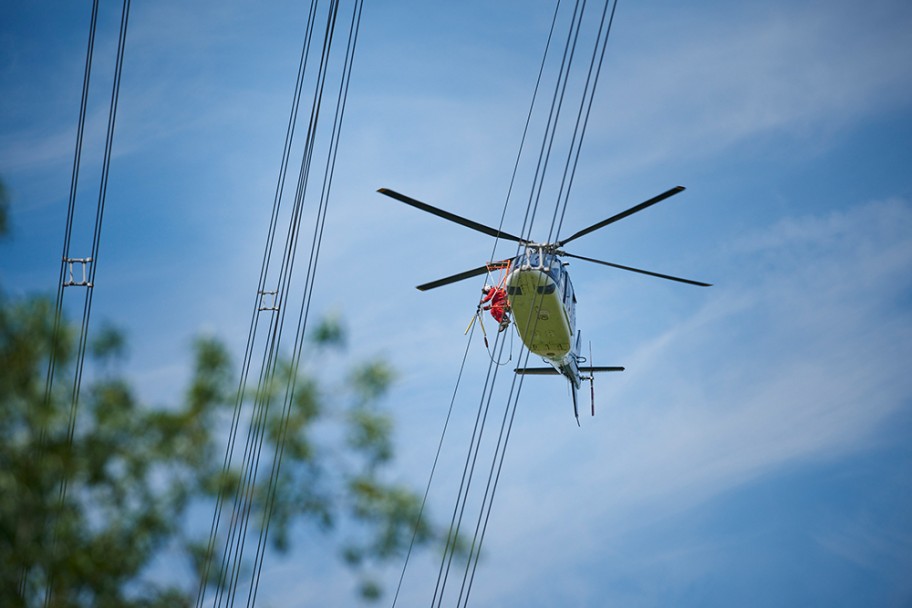 Das Foto aus der Froschperspektive zeigt einen unmittelbar neben der Leitung fliegenden, vierblättrigen Hubschrauber mit eingezogenem Fahrwerk. In der Tür des blauen Helikopters sitzt ein Monteur in rotem Overall, welche Vogelschutzmarkierungen anbringt.