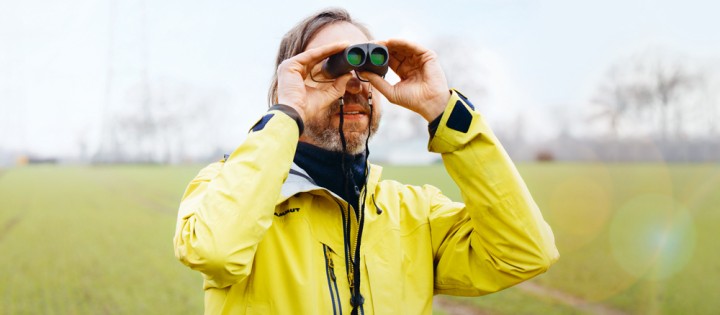 Das Bild zeigt einen Mann mit gelber Jacke, der durch ein Fernglas schaut.