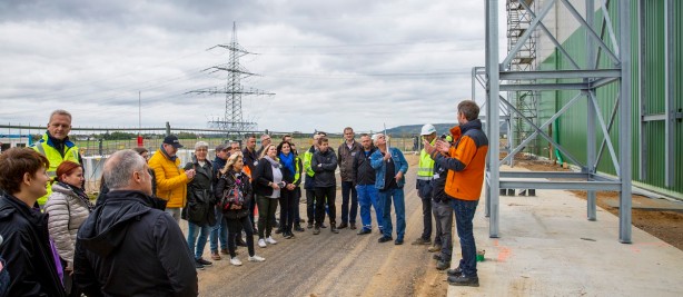 Das Bild zeigt eine Gruppe von Menschen auf einer Baustelle oder einem Industriegelände während einer Besichtigung. Ein Mann mit oranger Jacke spricht und gestikuliert, während die Zuhörenden aufmerksam lauschen. Einige der Personen tragen Warnwesten und Sicherheitskleidung, darunter auch ein Mann mit einem weißen Schutzhelm. Im Hintergrund sind ein hoher Strommast, ein Zaun und offene Landschaft zu sehen. Rechts im Bild befindet sich eine große grüne Industriehalle mit einem Stahlgerüst. Der Himmel ist bewölkt. Die Szene vermittelt den Eindruck einer Führung oder technischen Erklärung vor Ort.