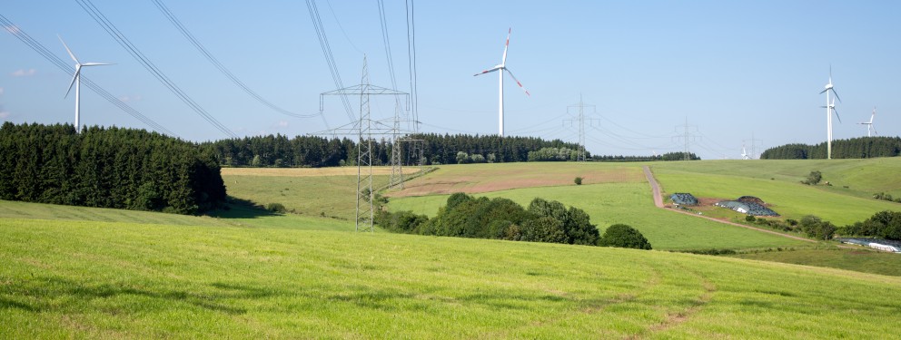 Das Bild zeigt eine Trasse in hügeliger Landschaft. Windräder sind am Horizont zu sehen.