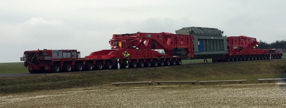 Das Bild zeigt einen Schwertransport auf einer Straße, der einen großen, massiven Transformator befördert. Der Transporter besteht aus mehreren rot lackierten Modulen mit zahlreichen Achsen und Rädern, um die Last zu verteilen. In der Mitte des Transports ist der grau-grüne Transformator sicher fixiert. Zwei Personen in Warnschutzkleidung und Helmen stehen vor dem Fahrzeug und prüfen offensichtlich den Transport. Der Hintergrund zeigt eine ländliche, leicht hügelige Landschaft unter einem bewölkten Himmel. 