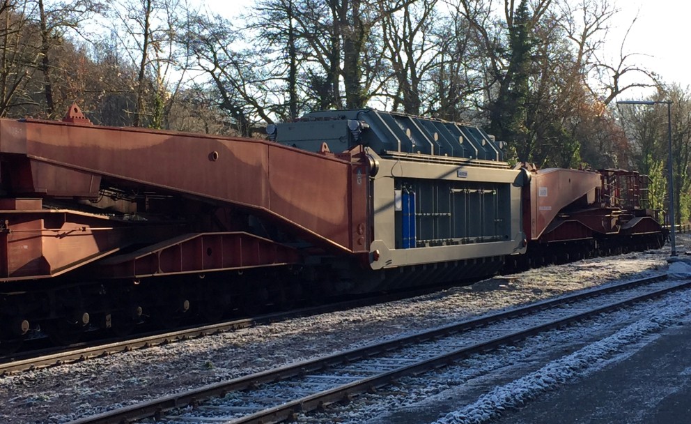 Das Bild zeigt einen großen Transformator, der auf einem speziellen Schwerlast-Eisenbahnwagen transportiert wird. Der Transformator ist grau-grün und massiv, sicher auf dem roten Transportgestell befestigt, das auf Schienen steht. Das Gestell besteht aus robusten Stahlträgern und zahlreichen Achsen, um das Gewicht zu verteilen. Die Umgebung zeigt eine winterliche Szene mit leichtem Schnee- oder Raureif auf den Bahnschwellen und Schienen. Im Hintergrund stehen kahle Bäume, was auf die kalte Jahreszeit hinweist.