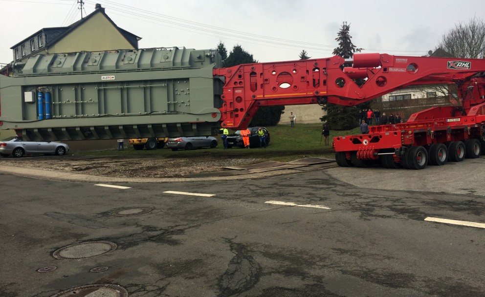Das Bild zeigt einen Schwertransport, der einen massiven, grau-grünen Transformator durch eine Kurve manövriert. Der Transformator ist auf einem roten, spezialisierten Transportfahrzeug mit vielen Achsen und Rädern befestigt, um das Gewicht zu verteilen. Mehrere Personen in Warnschutzkleidung stehen daneben und überwachen den Transport. Im Hintergrund befinden sich Wohnhäuser, parkende Autos und einige Schaulustige, die das Ereignis beobachten. Die Straße ist eng und zeigt deutliche Fahrspuren und Abdeckungen von Kanaldeckeln. Die Szene verdeutlicht die logistische Herausforderung und Präzision, die für den Transport solch großer Lasten in bebauten Gebieten erforderlich ist.