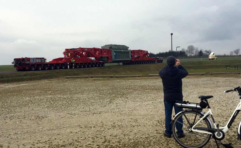 Das Bild zeigt einen Schwertransport mit einem großen, grau-grünen Transformator, der auf einem roten Spezialtransporter über eine schmale Straße fährt. Der Transporter besteht aus mehreren Modulen mit zahlreichen Achsen und Rädern. Zwei Personen in Warnschutzkleidung stehen an dem Fahrzeug und überwachen die Fahrt. Im Vordergrund fotografiert ein Mann in dunkler Kleidung die Szene, neben ihm steht ein weißes Fahrrad mit einem Akkuantrieb. Der Hintergrund zeigt eine offene, ländliche Landschaft mit einer Wiese und einem grauen, bewölkten Himmel. Ein kleines Fahrzeug, möglicherweise ein Begleitfahrzeug, steht weiter entfernt auf dem Feld. Die Atmosphäre wirkt ruhig und zeigt die Besonderheit des Transports in der Umgebung.