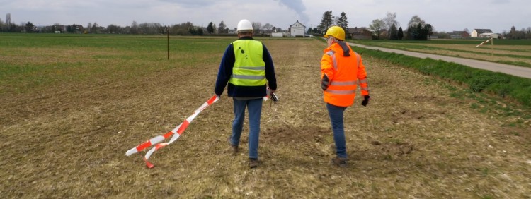 Das Bild zeigt zwei Menschen auf einem Feld. Einer der Menschen hält Flatterband in seinen Händen.