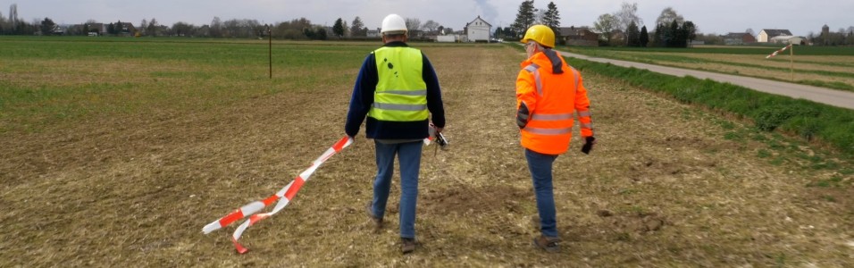 Das Bild zeigt zwei Menschen auf einem Feld. Einer der Menschen hält Flatterband in seinen Händen.