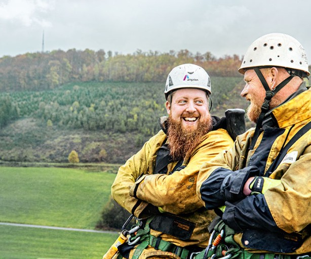 Das Bild zeigt zwei Männer in Sicherheitskleidung, bestehend aus gelben Jacken, Gurten und weißen Schutzhelmen mit Amprion-Logo, die sich fröhlich unterhalten. Der Mann links hat einen auffälligen roten Bart und lacht, während der Mann rechts ihm lächelnd zuhört. Im Hintergrund erstreckt sich eine hügelige Landschaft mit herbstlich gefärbten Bäumen und einer breiten, offenen Wiese. Auf der rechten Seite ist ein grüner, metallischer Teil einer Struktur sichtbar, der auf Arbeiten in luftiger Höhe hinweist, möglicherweise an Stromleitungen oder Masten