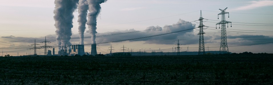 Dieses eher dunkel gehaltene Foto im Querformat zeigt den Blick über einen abgeernteten Acker auf ein Kraftwerk. Aus den Kühltürmen steigt Dampf auf. Im Vordergrund des Kraftwerks eine Stromleitung. 