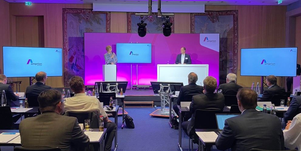 Das Bild zeigt eine klassische Vortragssituation. Zwei Menschen stehen auf der Bühne. Im Vordergrund sitzen etwa 20 Personen. Diese blicken auf die Bühne. Das Bild ist insgesamt im Lila getaucht. 