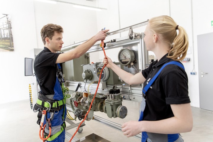 Zwei Azubis arbeiten am Umspannanlagen-Nachbau in Lotte. Einer trägt Klettergeschirr und befestigt ein orangenes Seil an der Anlage.